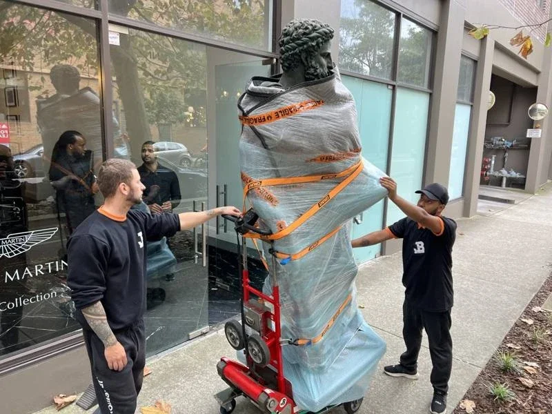 JB removalists with a wrapped bronze sculpture strapped to red powered moving equipment outside a showroom